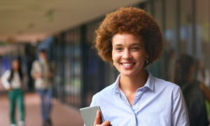 mulher negra com cabelo black power ruivo, camisa de botão azul segurando um tablet, em um ambiente aberto de escola.