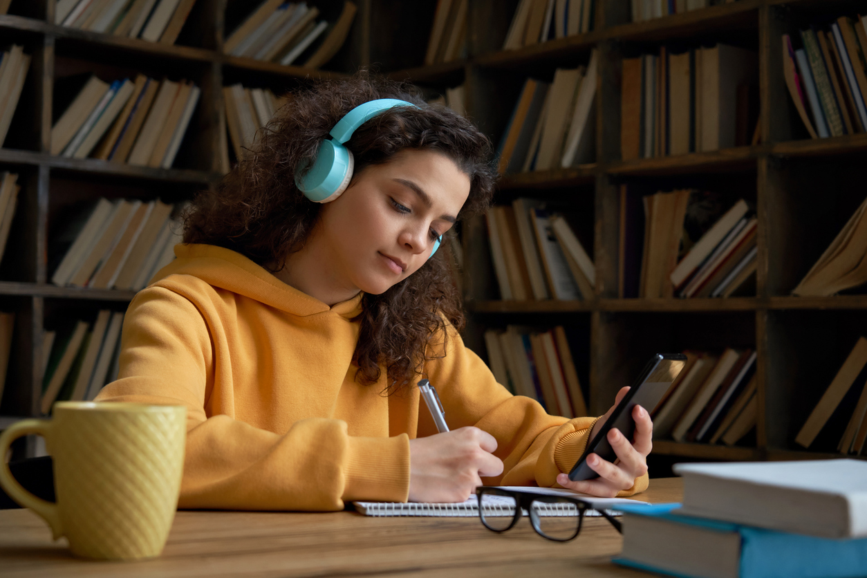 Jovem do ensino médio observa o celular enquanto faz anotações.